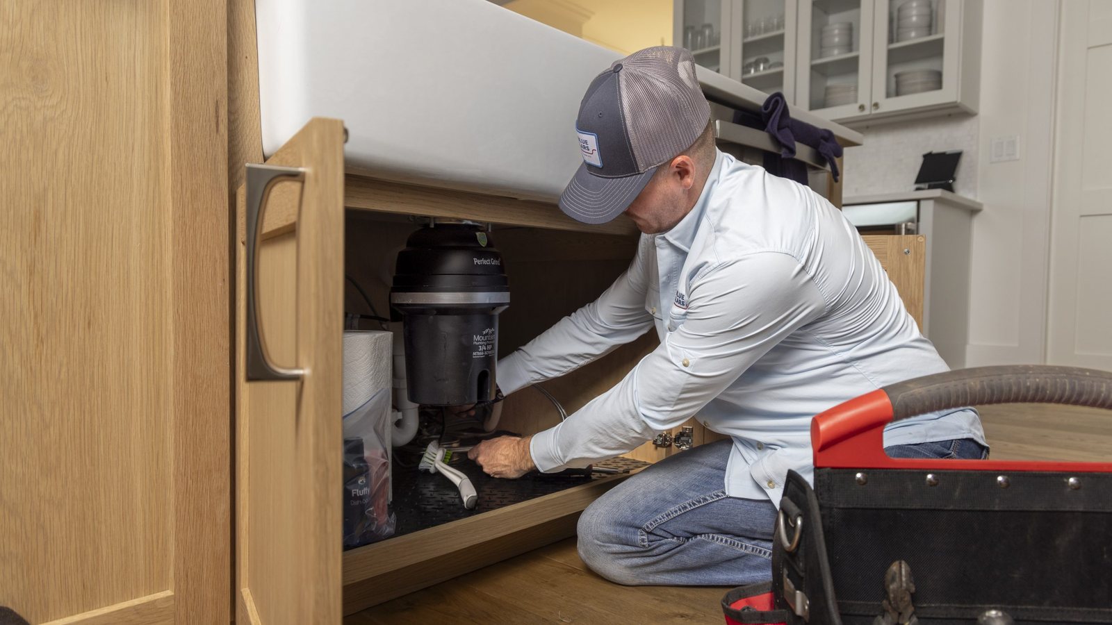 Blue Collars technician kneeling at an open kitchen cabinet working on the drain beneath a sink, with a tool bag beside him