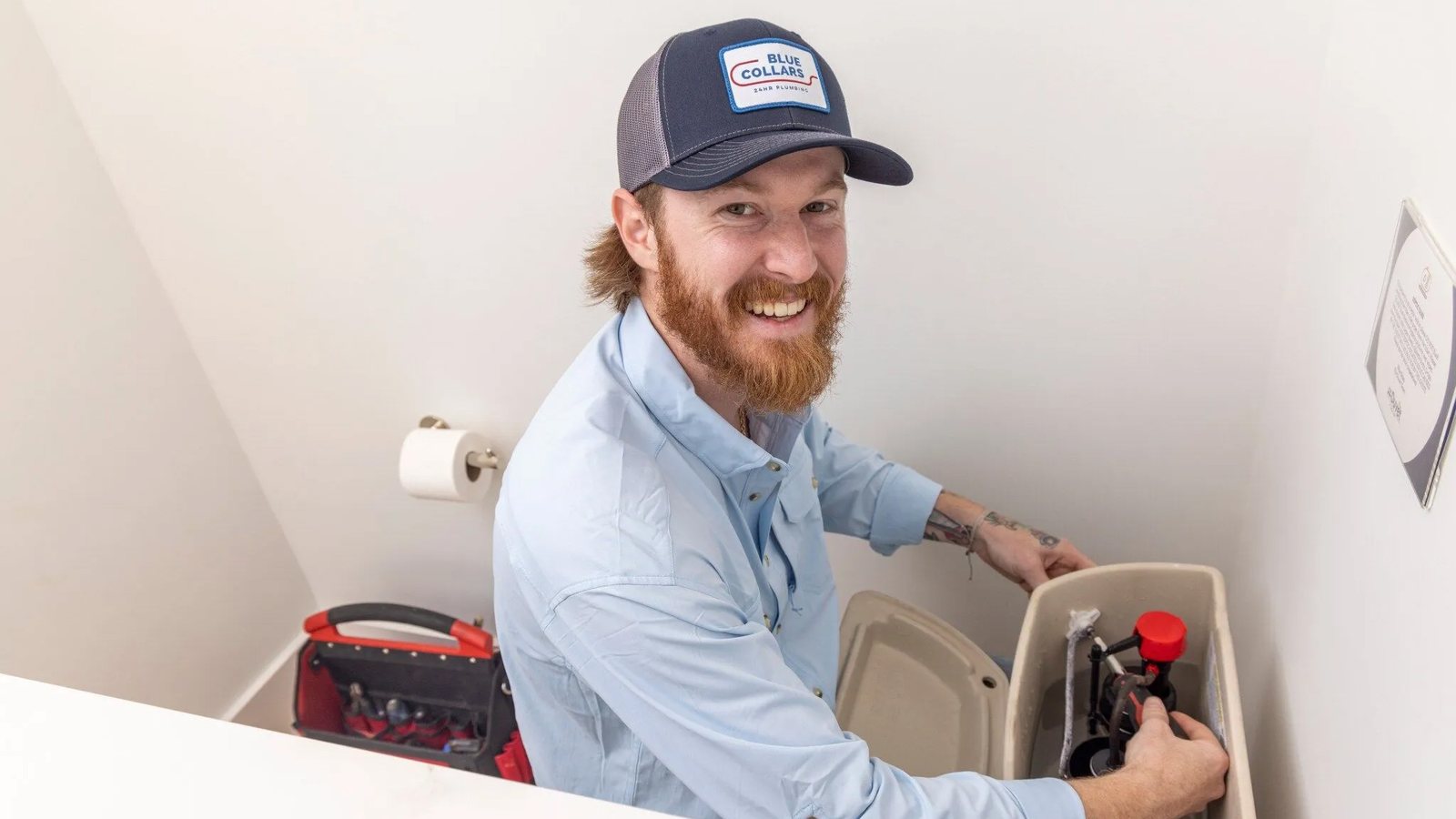 Blue Collars technician reaching into an open toilet tank to check the fill valve, with a tool bag on the floor behind him