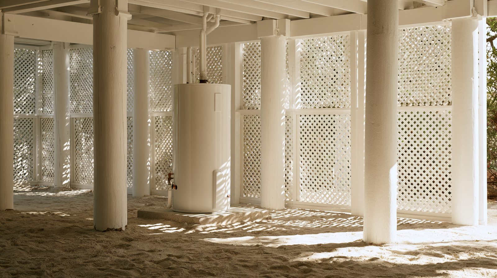 Residential water heater standing on a concrete pad in the open ground level area beneath an elevated Lowcountry house, with white piles and lattice screening